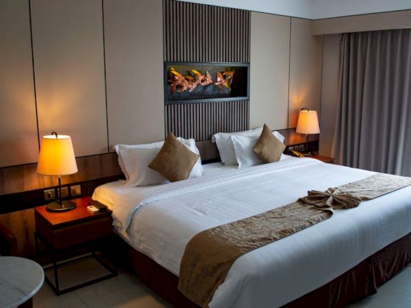A modern hotel room with a large bed, two bedside tables with lamps, cushioned headboard, a wall-mounted artwork, and curtains drawn to one side.