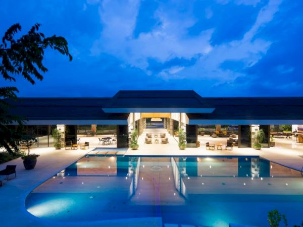 A luxurious, modern building with a large outdoor swimming pool illuminated at dusk, featuring open spaces and seating areas.