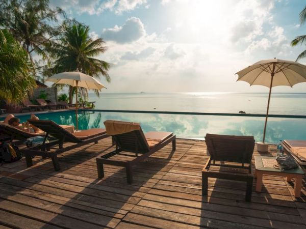 A serene poolside scene with lounge chairs and umbrellas overlooking a calm ocean, framed by palm trees, under a bright, cloudy sky.