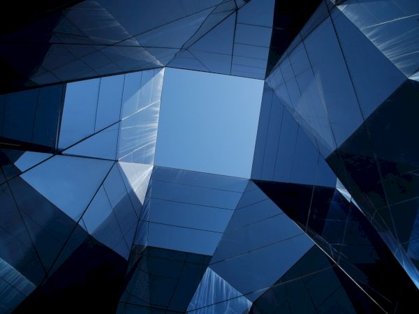 This is an upward view of a geometric structure made of reflective glass panels against a blue sky, forming an abstract architectural pattern.