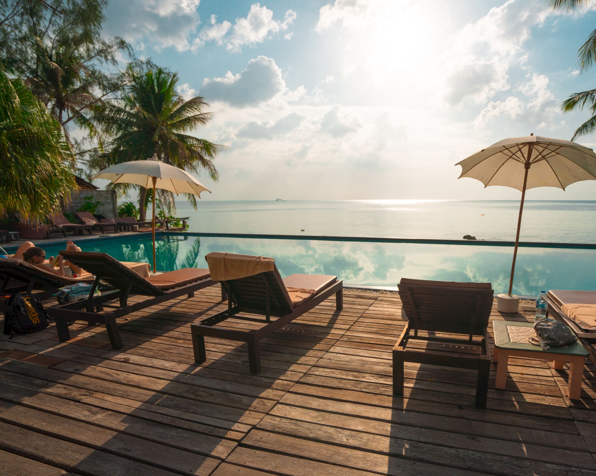 A serene poolside scene with lounge chairs, umbrellas, and palm trees overlooking the ocean under a partly cloudy sky during daylight hours.