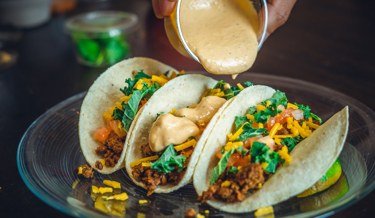 Three tacos filled with ground meat, shredded cheese, tomatoes, and greens are being topped with a creamy sauce from a small container.
