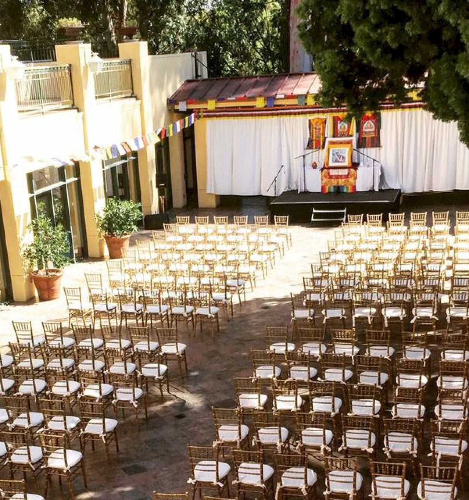 The image shows an outdoor venue set up for an event, with rows of chairs facing a stage adorned with decorations, surrounded by trees and buildings.