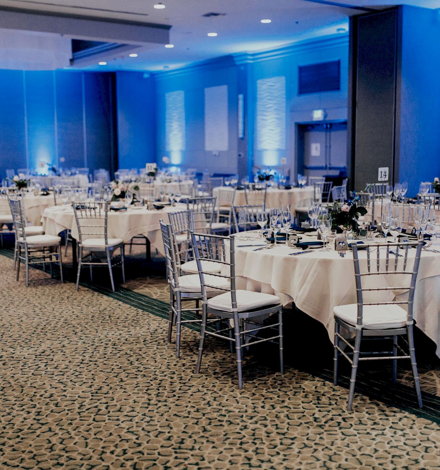 The image shows a banquet hall set up with round tables covered in white tablecloths, silver chairs, and blue lighting, ready for an event.