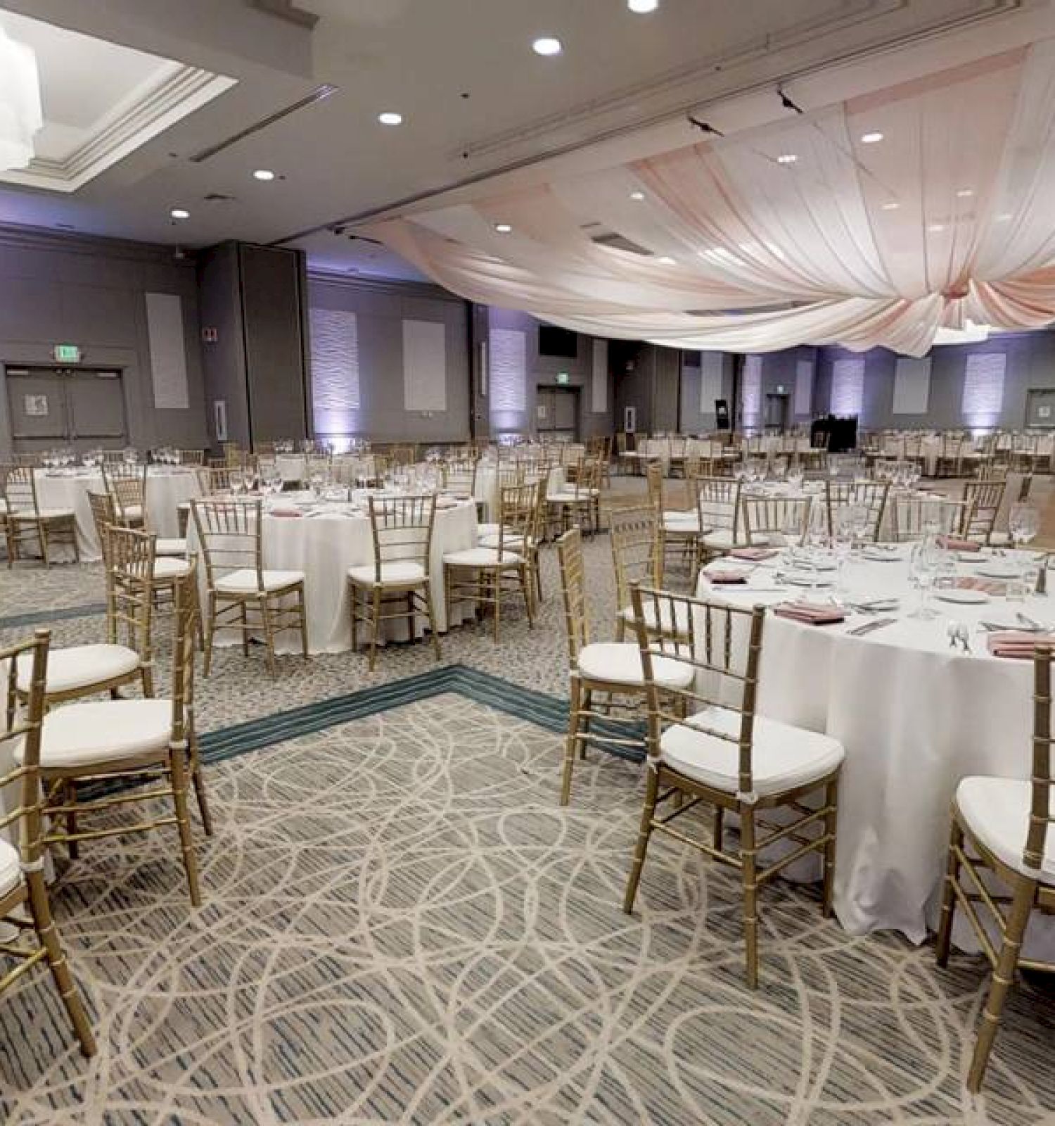 A large, elegantly decorated banquet hall with round tables, gold chairs, and white tablecloths, set up for an event with draped ceiling fabric.