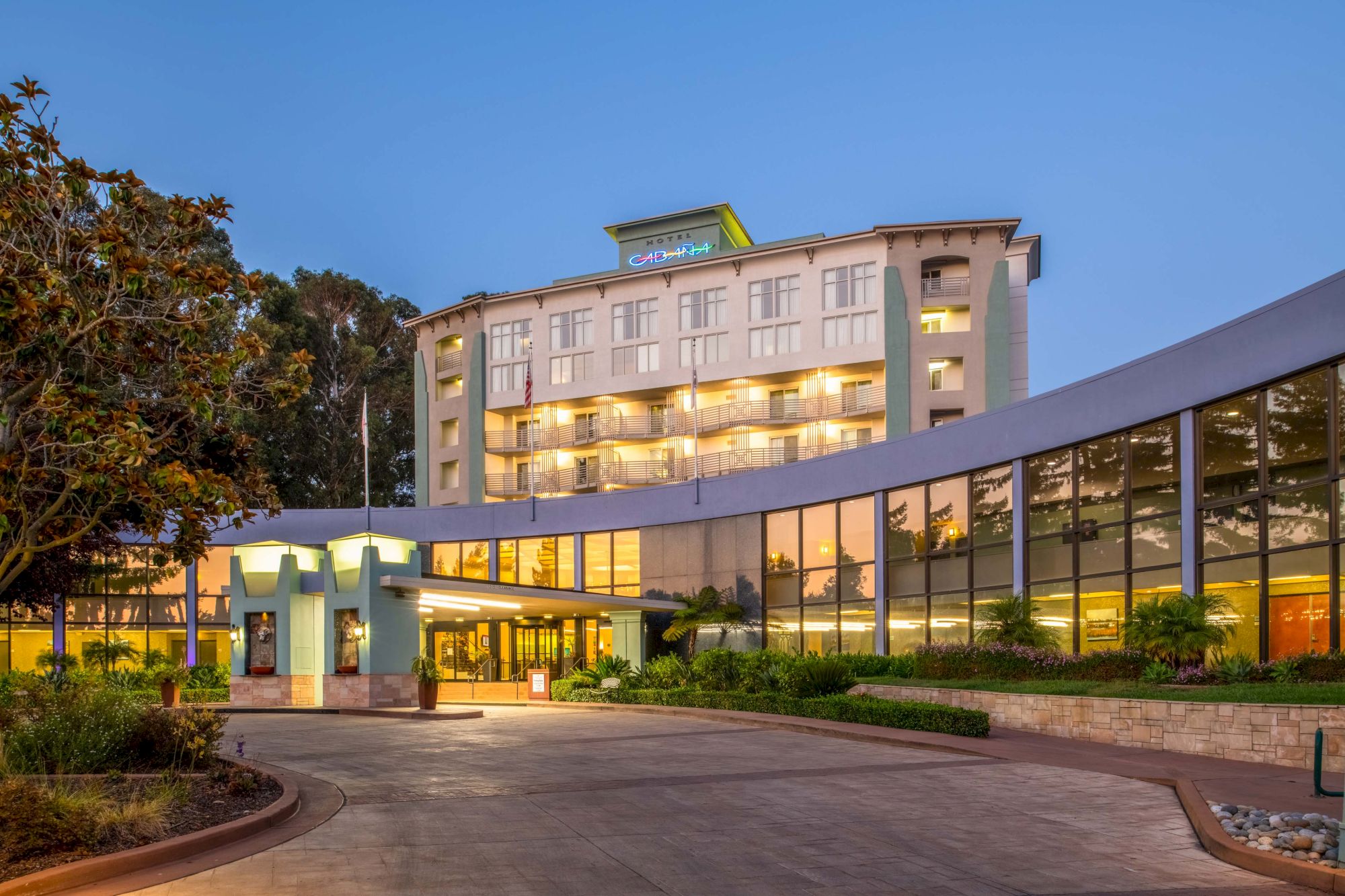 The image shows the exterior of a multi-story hotel building with large windows and a curving driveway at dusk, surrounded by greenery and trees.