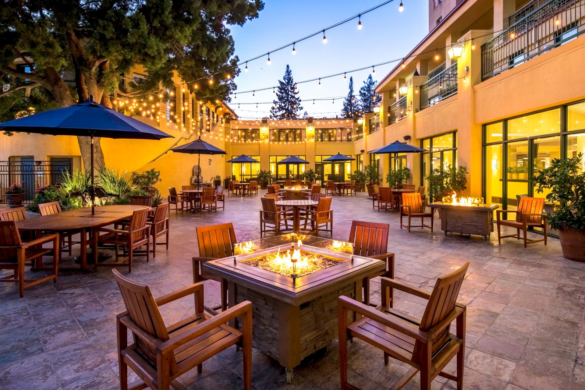 An outdoor patio with wooden tables and chairs, fire pits, string lights, and surrounding buildings, creating a cozy and inviting atmosphere.
