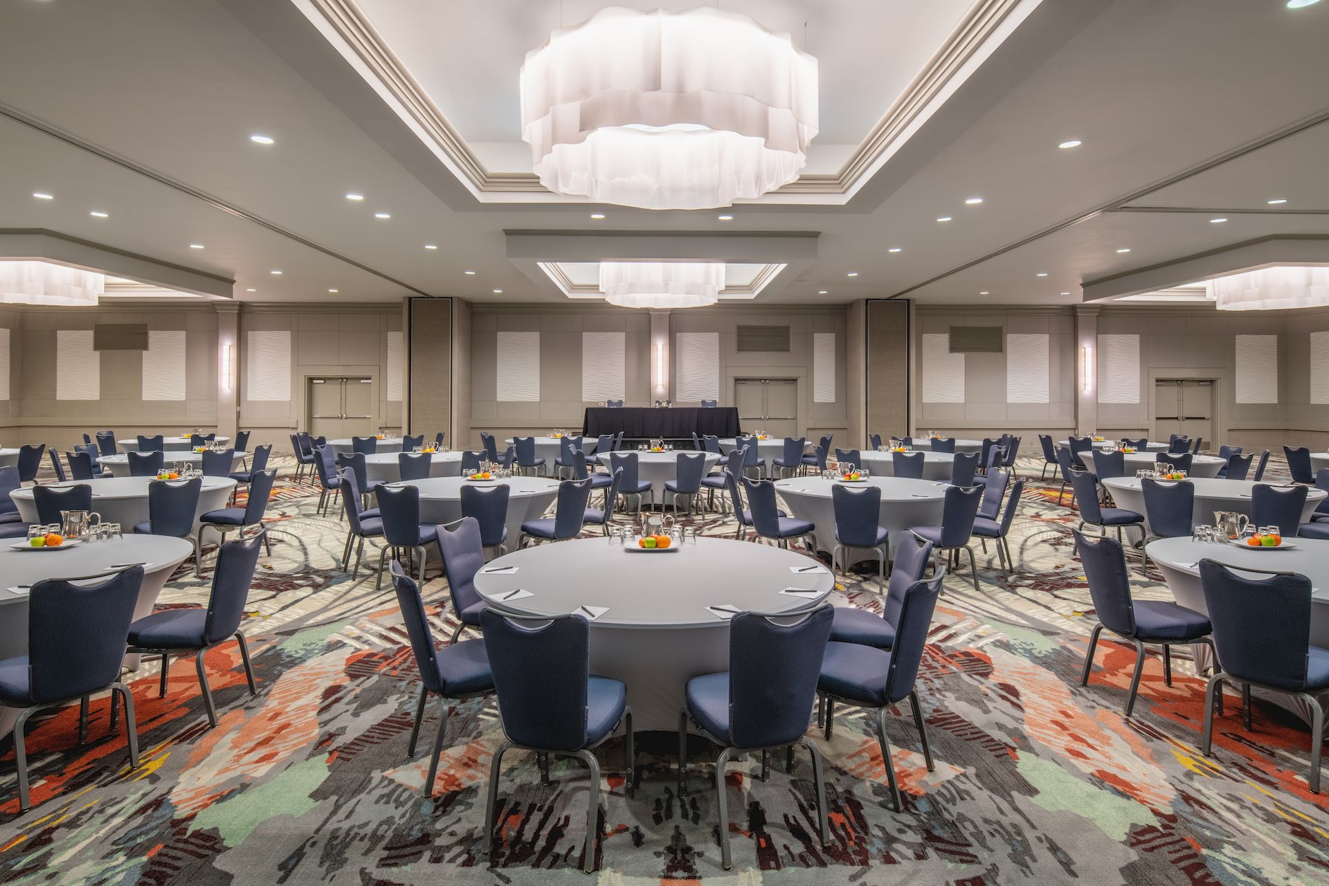 The image shows a large, modern conference room set up with round tables, blue chairs, colorful carpet, and elegant ceiling lights.