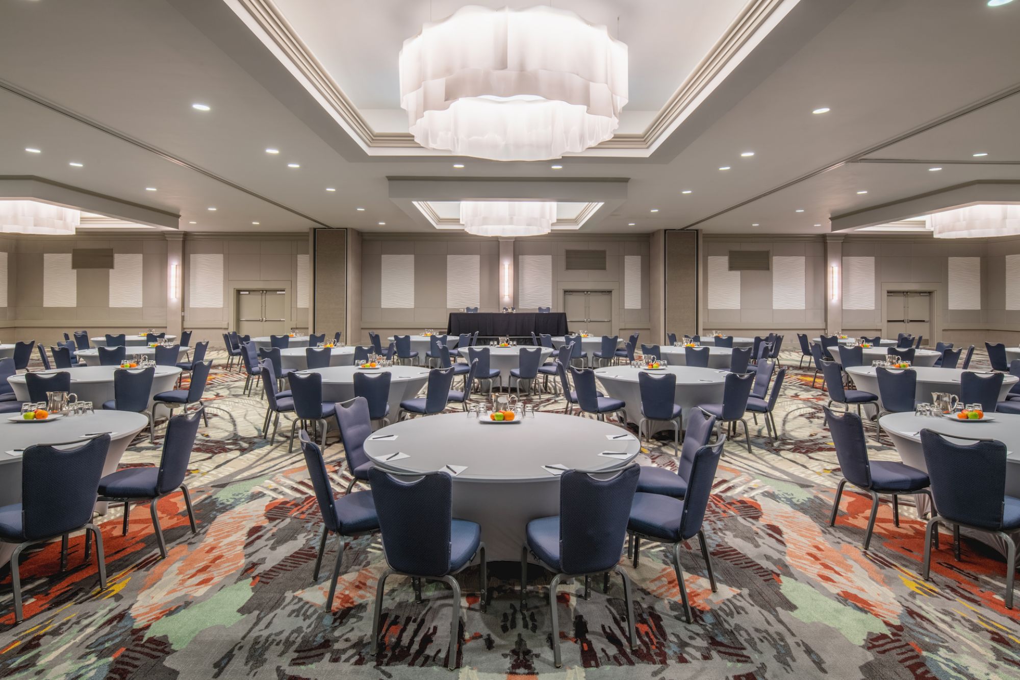 The image shows a large, modern conference room set up with round tables, blue chairs, colorful carpet, and elegant ceiling lights.