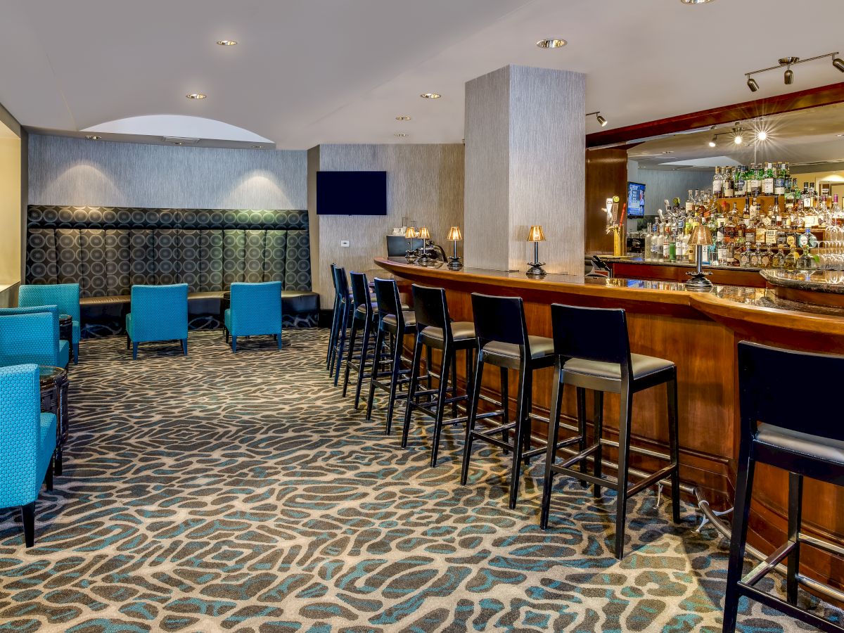 This image shows a bar with high stools, a fully stocked bar counter, and a seating area with blue armchairs and a patterned carpet.