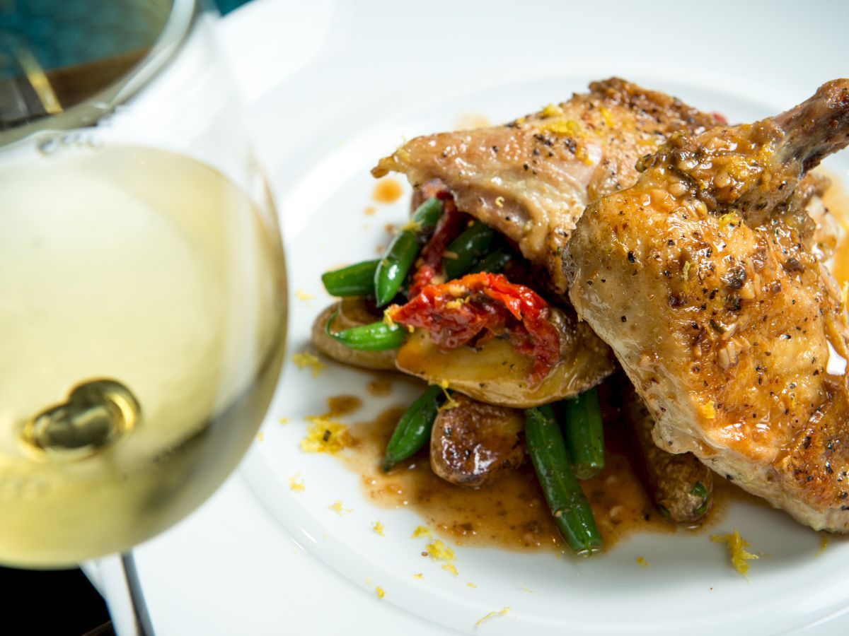 A deliciously prepared chicken dish with vegetables and a glass of white wine, presented on a white plate and photographed up close.
