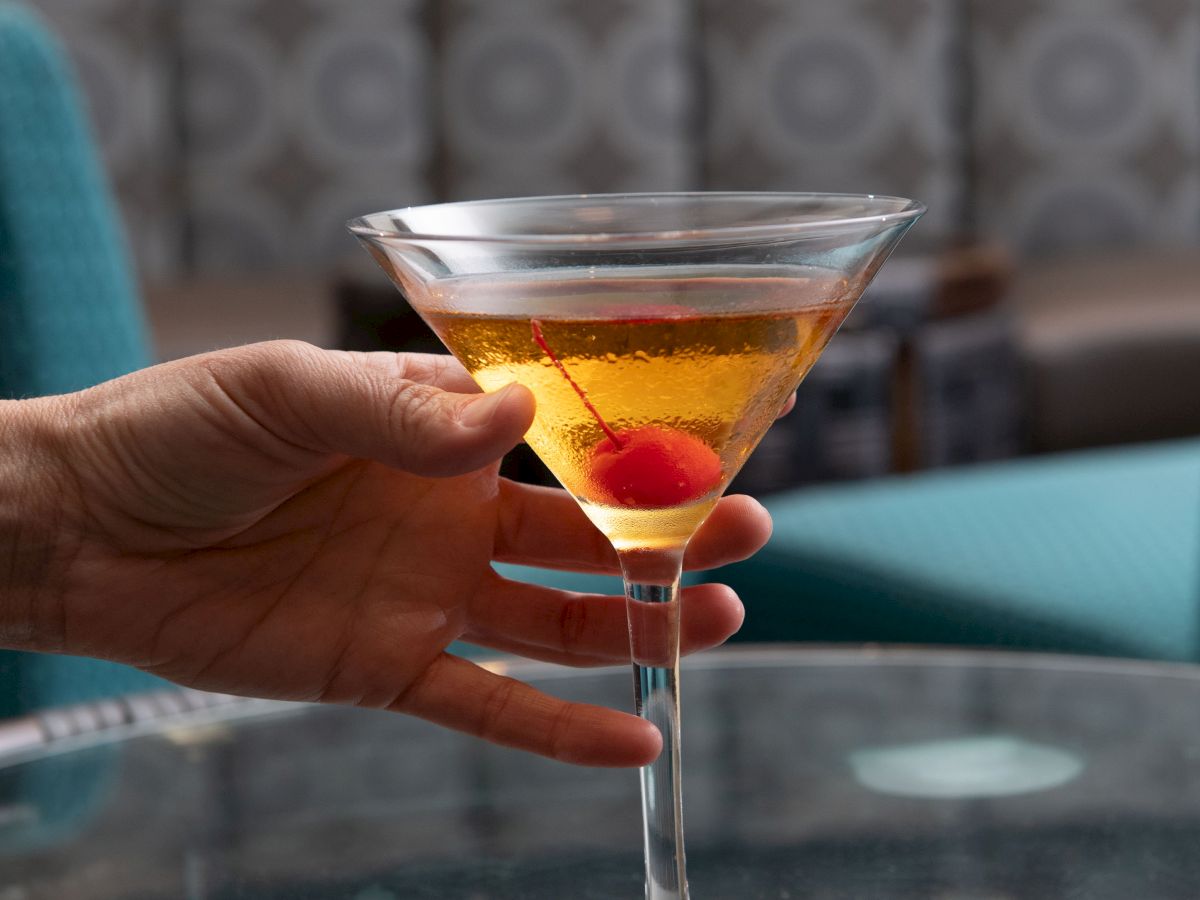 A hand is holding a martini glass containing a golden drink with a cherry inside, over a glass table, with teal furniture in the background.