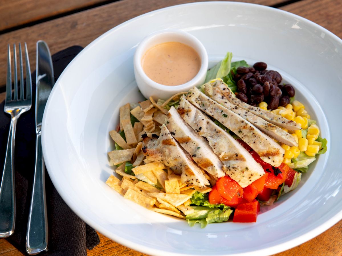 A bowl filled with sliced grilled chicken, diced tomatoes, corn, black beans, tortilla strips, and a small cup of dressing, next to a fork and knife.