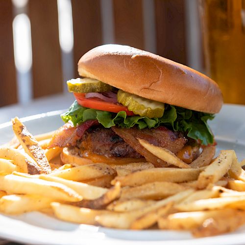 A plate of French fries with a cheeseburger topped with lettuce, tomato, pickles, and bacon, with a glass of beer in the background.