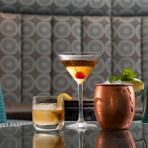 Three cocktails sit on a glass table: a whiskey drink with lemon peel, a martini with a cherry, and a copper mug drink with mint garnish.