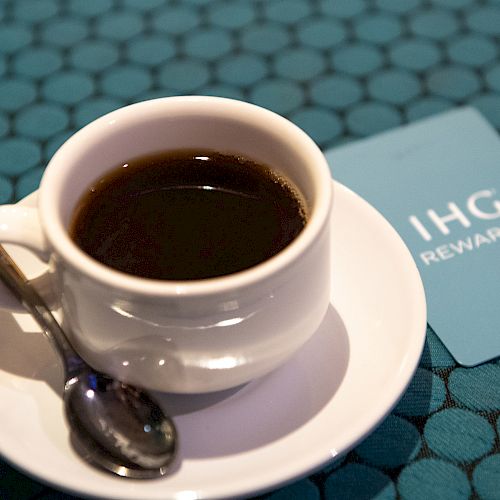 A cup of black coffee on a saucer with a spoon, next to an IHG Rewards card, set on a teal-patterned tablecloth.