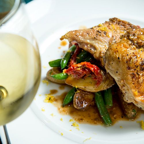 The image shows a gourmet dish with cooked poultry, green beans, potatoes, and a white sauce, accompanied by a glass of white wine on the side.