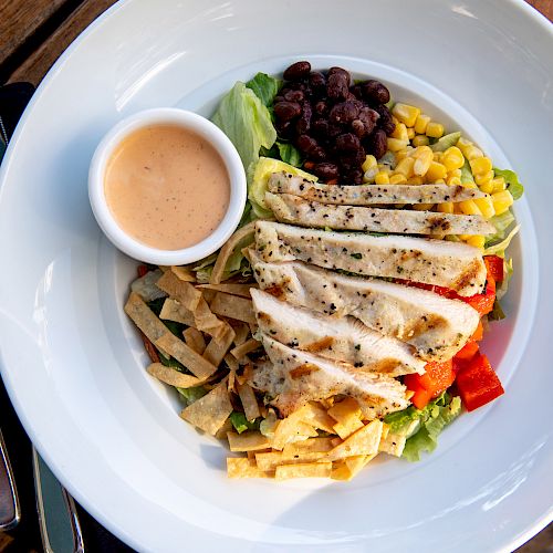 A mixed salad with grilled chicken strips, black beans, corn, tortilla strips, and dressing on the side is on a white plate with cutlery nearby.