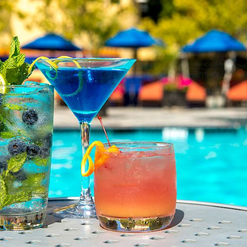 The image shows three colorful cocktails placed on a table by a poolside, with blue umbrellas and lounge chairs in the background.