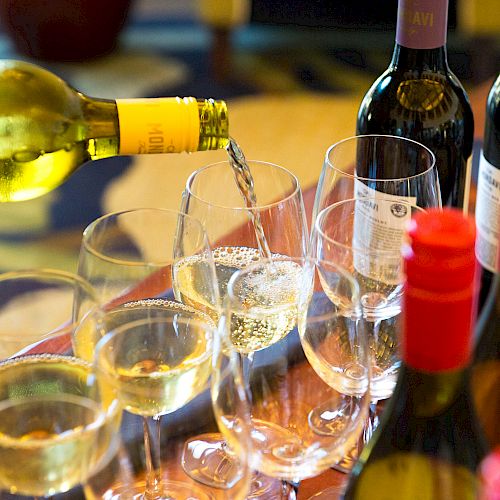 A hand is pouring white wine into a glass with several other wine glasses and bottles of red and white wine on a table.