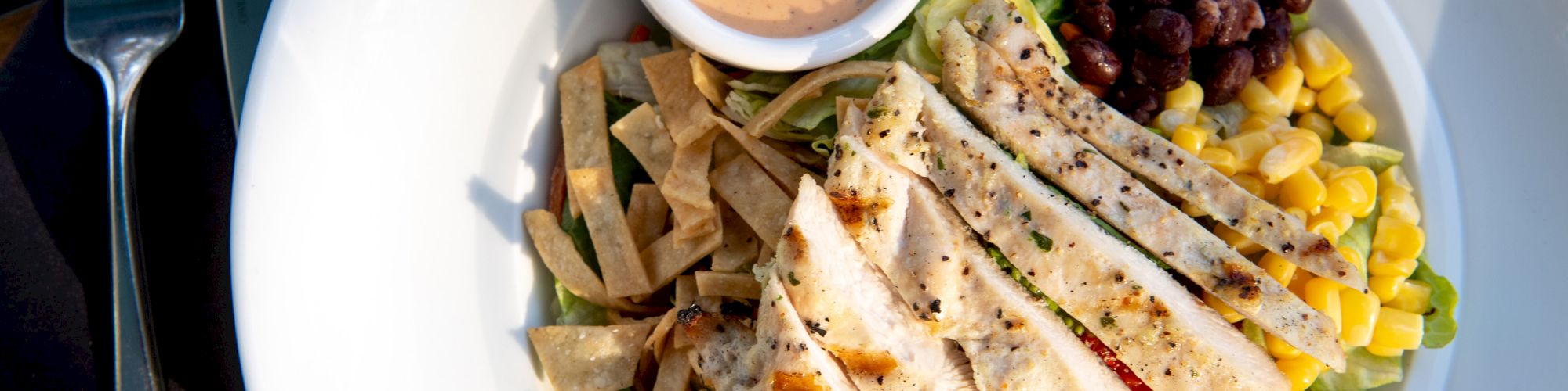 The image shows a salad with grilled chicken, black beans, corn, tortilla strips, red bell peppers, and a cup of dressing, served in a white bowl.