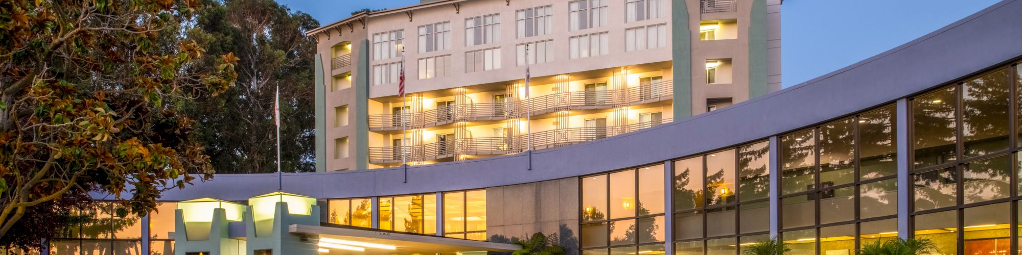 The image shows a modern multi-story hotel with a curved glass facade and a driveway entry, surrounded by greenery, in an evening setting.