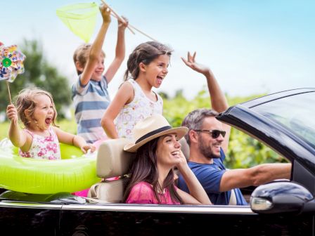 A family of five is enjoying a drive in a convertible car, with the children excitedly playing, one with a floatie and another holding a fishing net.