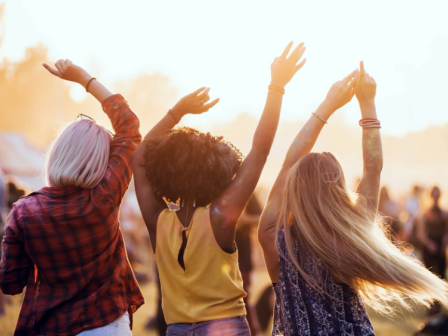 Three people are dancing outdoors with their arms raised, enjoying a sunny day at what appears to be a festival or concert, bathed in natural light.