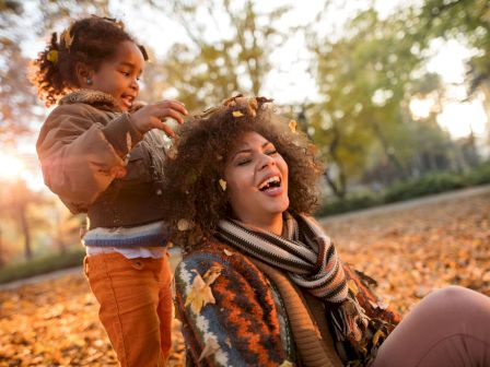 A child places autumn leaves on a woman's head as they both laugh and enjoy a beautiful fall day in a park covered with leaves, illuminated by sunset.