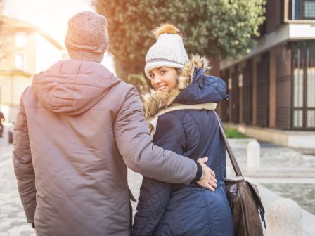 A couple in winter attire, walking outdoors. The woman smiles at the camera while the man has his arm around her.