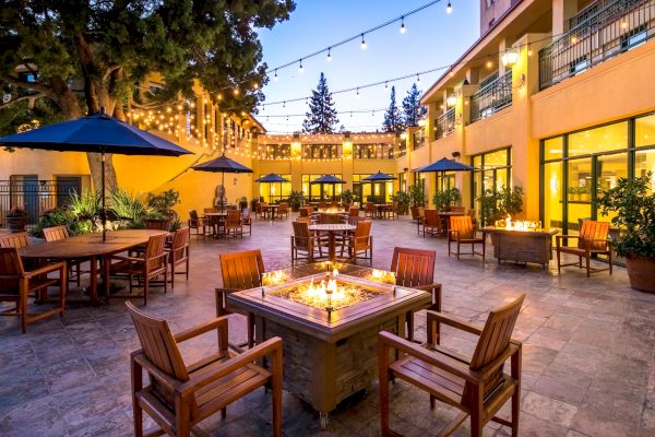 An outdoor patio with string lights features tables with chairs, umbrellas, and fire pit seating, surrounded by a warm and inviting ambiance.