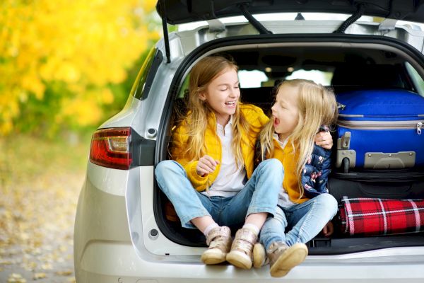 Two young children are sitting in the open trunk of a car, laughing together. The trunk is packed with suitcases and a blanket.