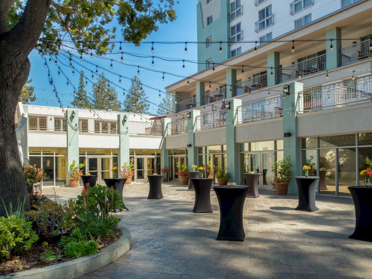 An outdoor patio area with string lights, cocktail tables with black covers, and potted plants, adjacent to a multi-story building.