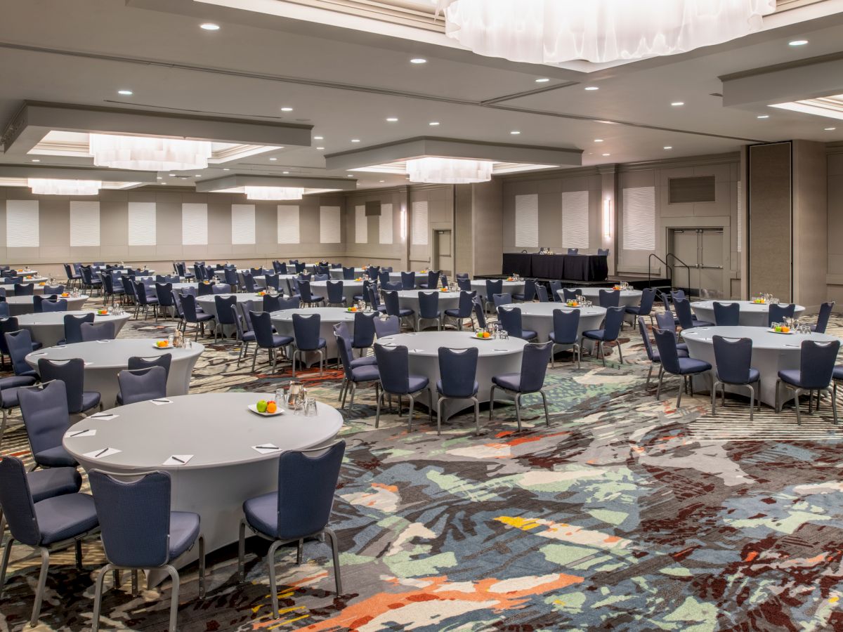 A banquet hall with round tables and blue chairs set up, featuring colorful patterned carpet and modern lighting fixtures, ending the sentence.