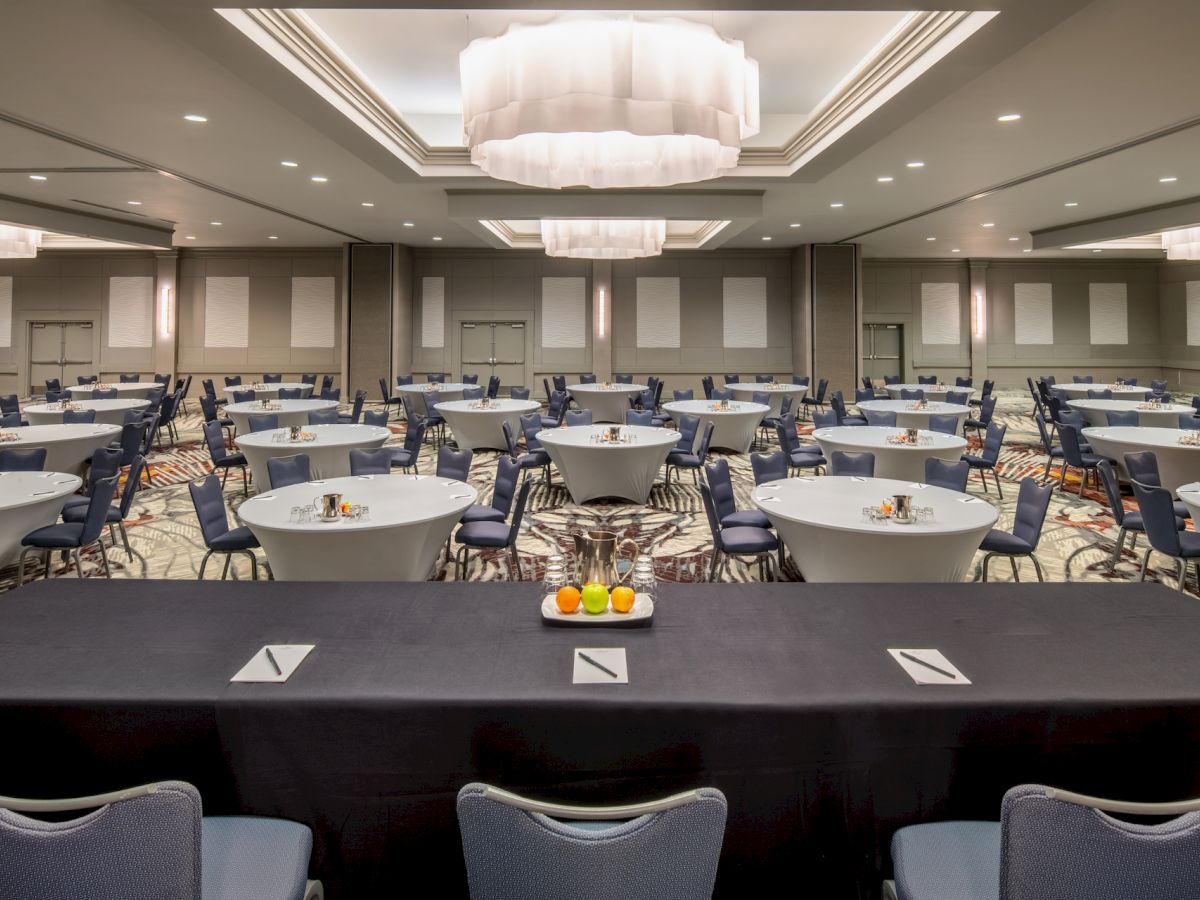 An empty conference room with round tables and chairs arranged for an event, viewed from behind a speaker's table adorned with fruits and notepads.