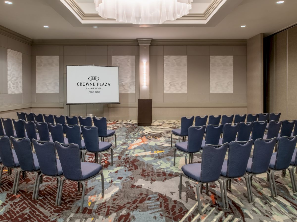 A conference room with rows of blue chairs facing a podium and projection screen showing the Crowne Plaza hotel logo.