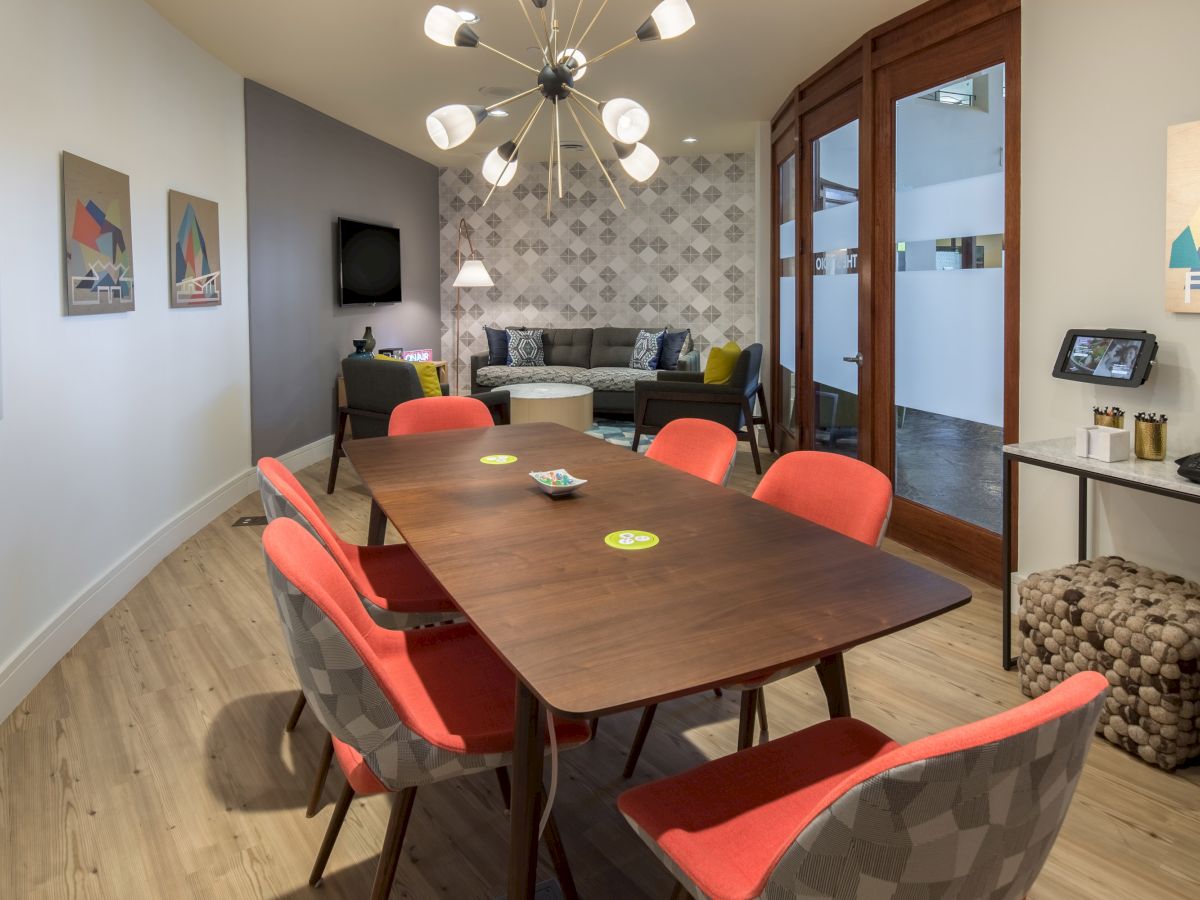 A modern meeting room with a wooden table, orange and gray chairs, wall art, a unique chandelier, and a seating area in the back.