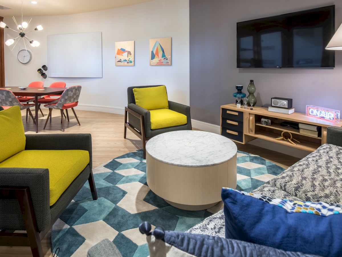 A modern living room with colorful furniture, a round coffee table, TV, and adjacent dining area with red chairs. Art decorates the walls.