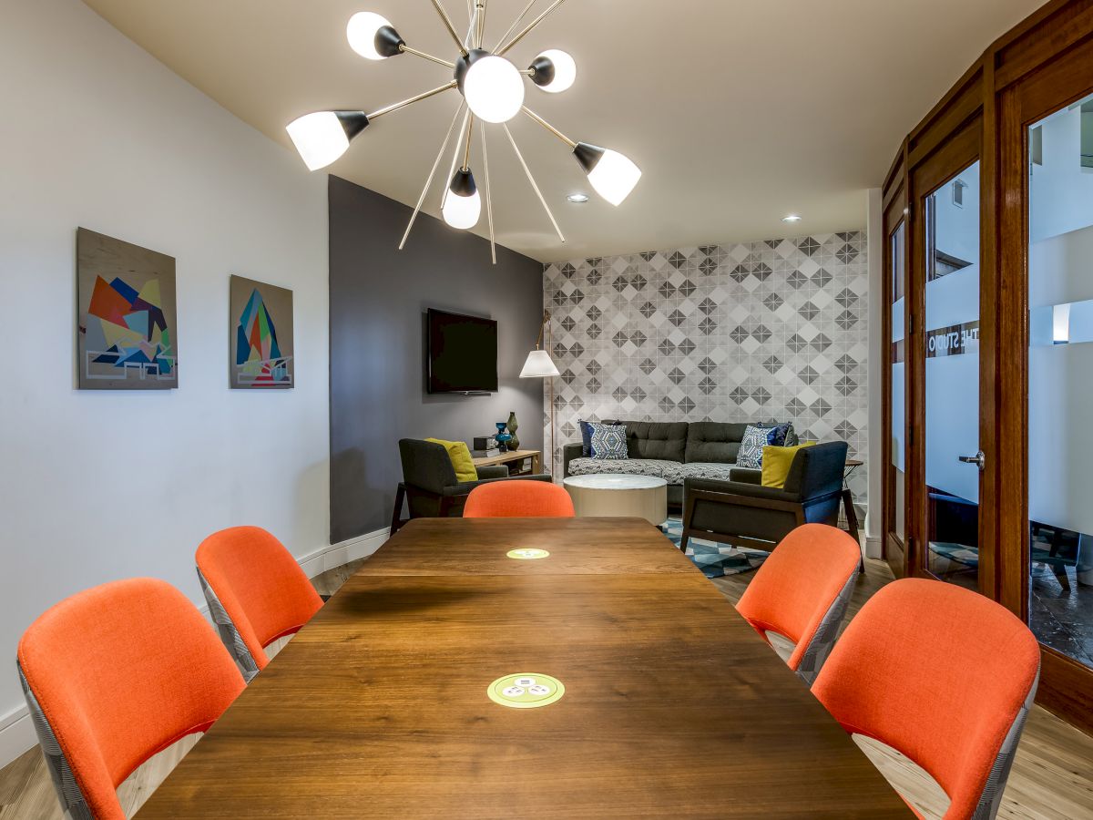 The image shows a conference room with a wooden table, orange chairs, a modern light fixture, abstract art on the walls, and a lounge area in the background.