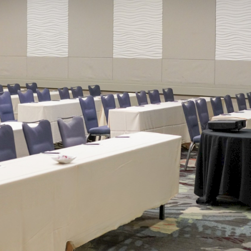 A conference room with rows of blue chairs and white-table desks, a black round table at the front, beige walls, and patterned carpet.