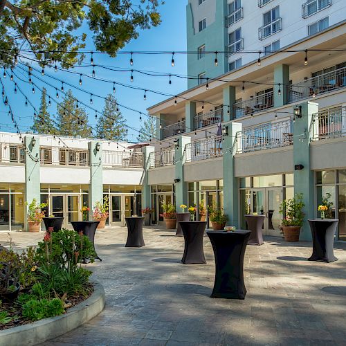 The image shows an outdoor patio area with tall cocktail tables, string lights, and potted plants. A multi-story building is in the background.