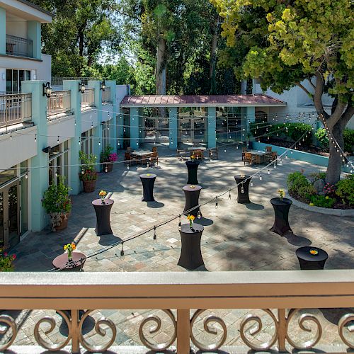 An outdoor event space with cocktail tables, decorations, and plenty of greenery, set up in a courtyard between buildings, ready for guests.