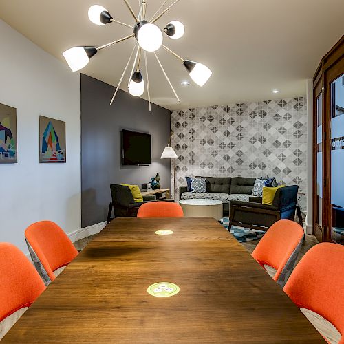 A modern conference room features a wooden table with orange chairs, a contemporary chandelier, artwork, TV, and a cozy lounge area.