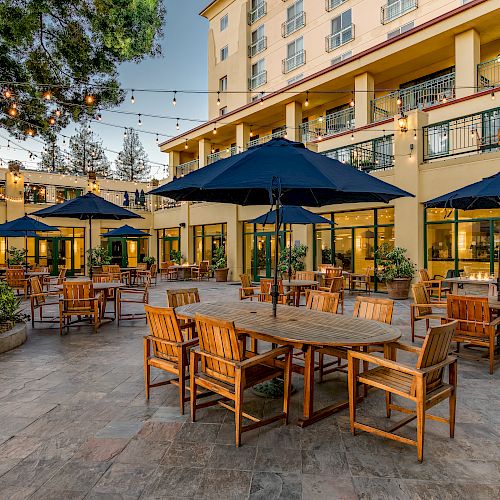 An outdoor dining area with wooden tables and chairs, blue umbrellas, string lights, surrounded by a multi-story building in the background with balconies.