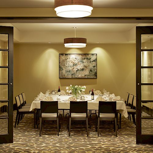 A dining room with a long table set for dinner, surrounded by chairs, with two pendant lights, a painting on the wall, and glass doors.