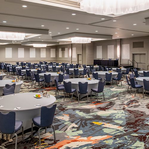 This image shows a large conference room with round tables, blue chairs, colorful carpet, and a stage at the front. The room is well-lit.