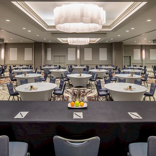 The image shows a conference room with rows of round tables and chairs arranged for an event, with a table at the front set for speakers.