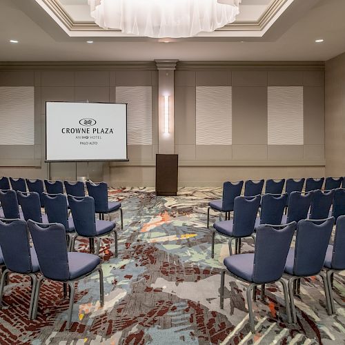 The image shows a conference room with rows of blue chairs facing a podium and a projection screen reading 