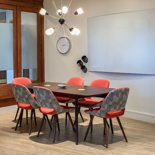 A modern conference room with a table, six chairs, a clock, and a contemporary light fixture. A whiteboard and abstract art decorate the walls.