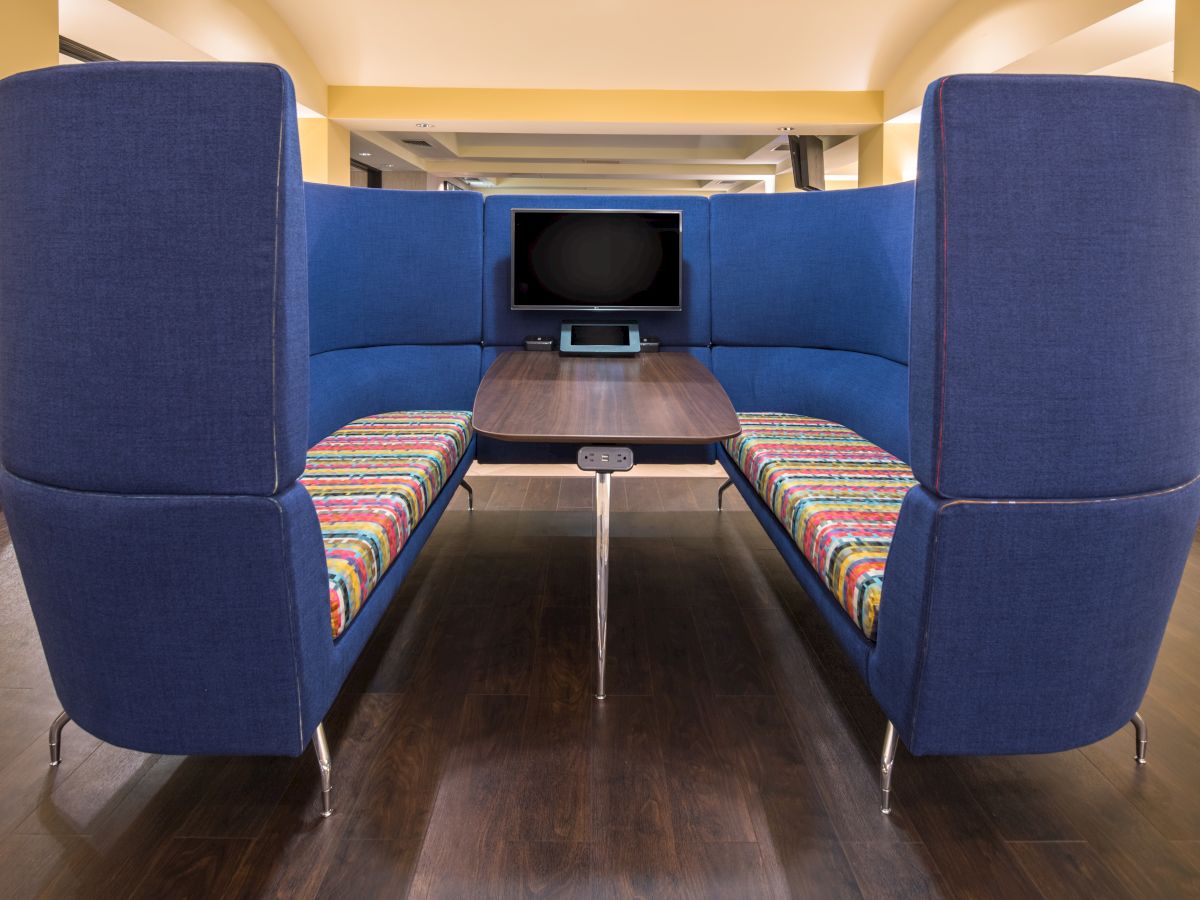 The image shows a modern seating area with blue high-back sofas, a wooden table, a mounted TV screen, and striped cushions in a brightly lit room.